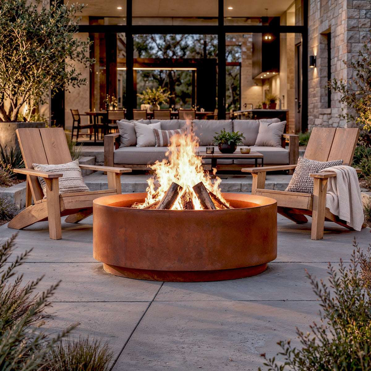 The Jake Corten steel fire pit with a wood fire between two wooden lounge chairs on a stone patio
